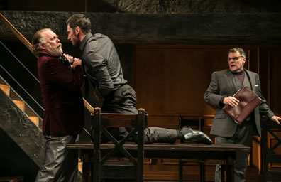 Richard in black shirt and pants kneeling on a table grabs the throat of Hastings standing at one end of the table--he in red dinner jacket and gray leather pants--as Stanley with briefcast and wearing a grey three-piece suit with turtleneck sweater shirt, looks on in consternation. The stairway and wood-paneled wall is int he background.